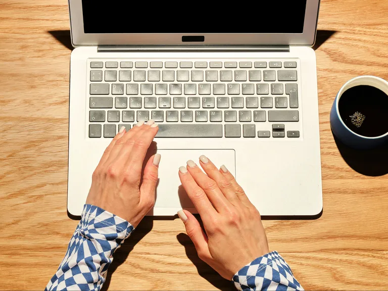 Hände tippen auf einer Laptop-Tastatur, daneben steht eine Tasse Kaffee auf einem Holztisch. Die Frau vergleicht online unterschiedliche Lohnmodelle.